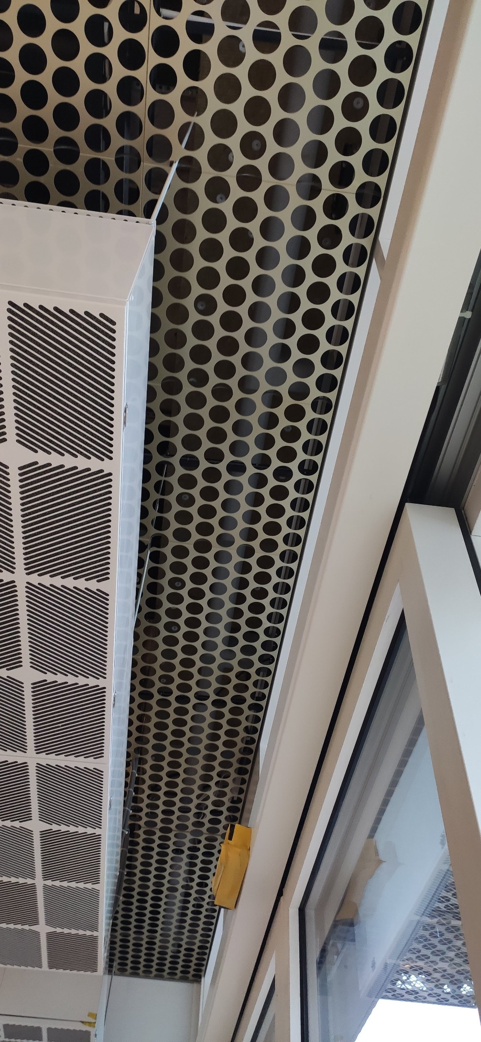 Metalowy sufit podwieszany z perforacją w postaci okręgów, sąsiadujący z kratką wentylacyjną i oknem, widziany pod kątem z dołu.