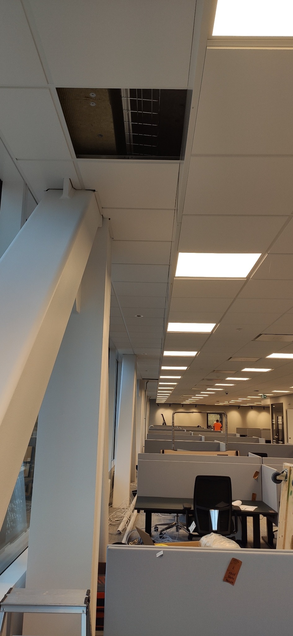 Otwór w kasetonowym suficie biurowym, widoczne elementy instalacji, białe kolumny konstrukcyjne, stanowiska pracy w tle.