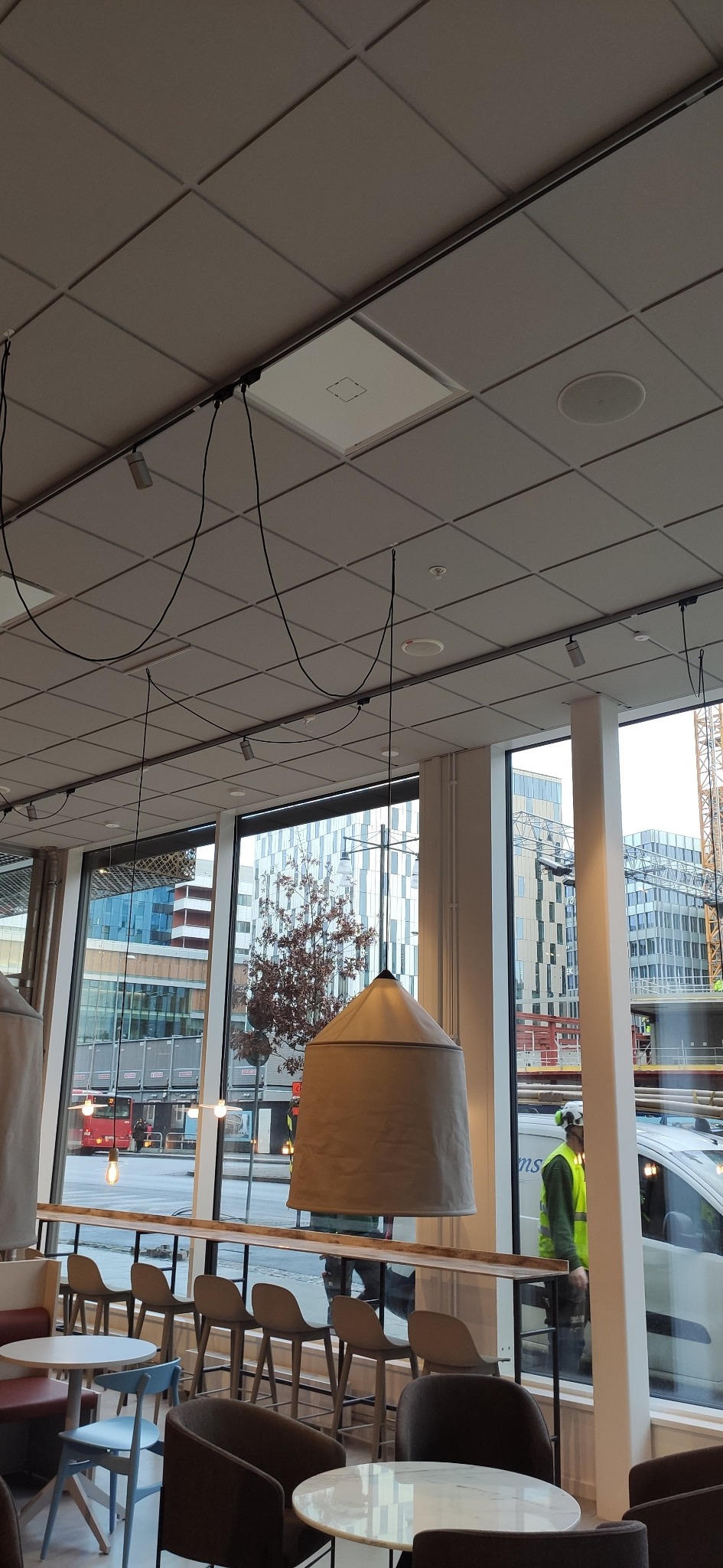 Sufit kasetonowy z zamontowanymi lampami punktowymi i wiszącymi lampami w kształcie stożka w lokalu biurowym z widokiem na budynki w tle.