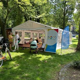 DREAM TENT SEBASTIAN MATYSZCZAK - Biały namiot eventowy w parku, z osobami w środku. Obok rower, flagi i tablice informacyjne. Słoneczny dzień, zielona trawa i drzewa w tle.