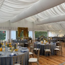 DREAM TENT SEBASTIAN MATYSZCZAK - Uroczyste przyjęcie w namiocie: okrągłe stoły z szarymi obrusami, złote krzesła, kwiatowe dekoracje i elegancka zastawa. Biały sufit z draperiami i żyrandol.