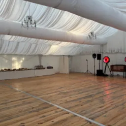 Elegancki namiot bankietowy z drewnianą podłogą, oświetleniem dekoracyjnym i stołem z przekąskami, gotowy na przyjęcie. Widoczne żyrandole i sprzęt nagłośnieniowy.