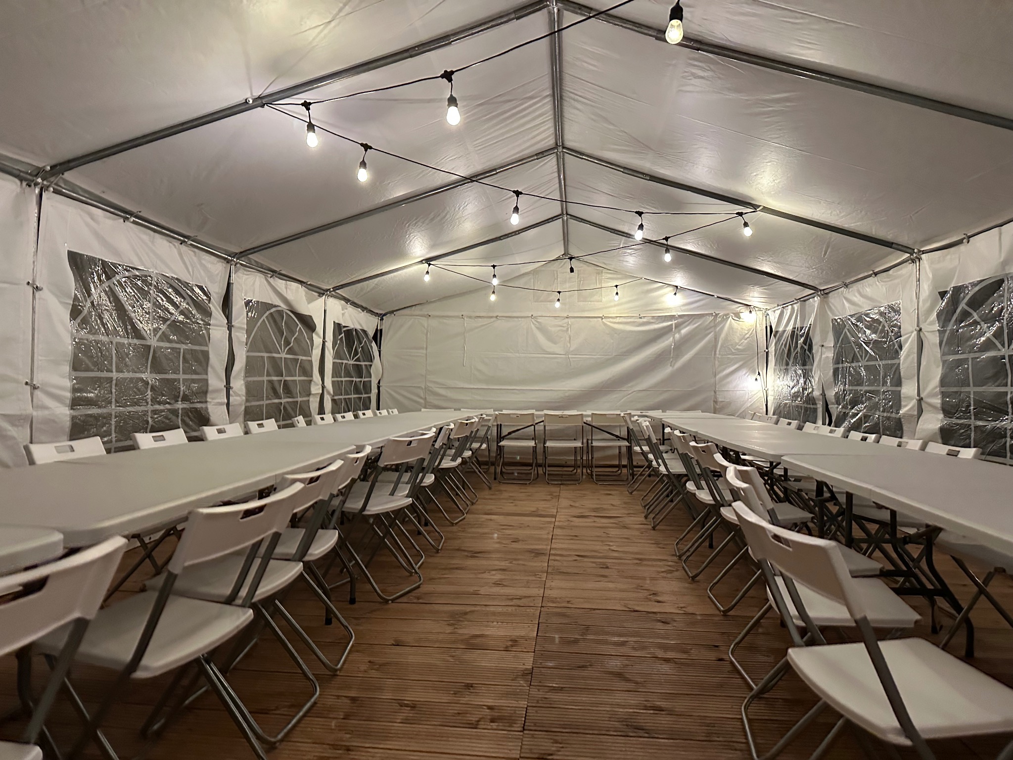 Wnętrze białego namiotu eventowego z drewnianą podłogą, oświetlonego girlandami żarówek. Ustawione białe stoły i krzesła, gotowe na przyjęcie.