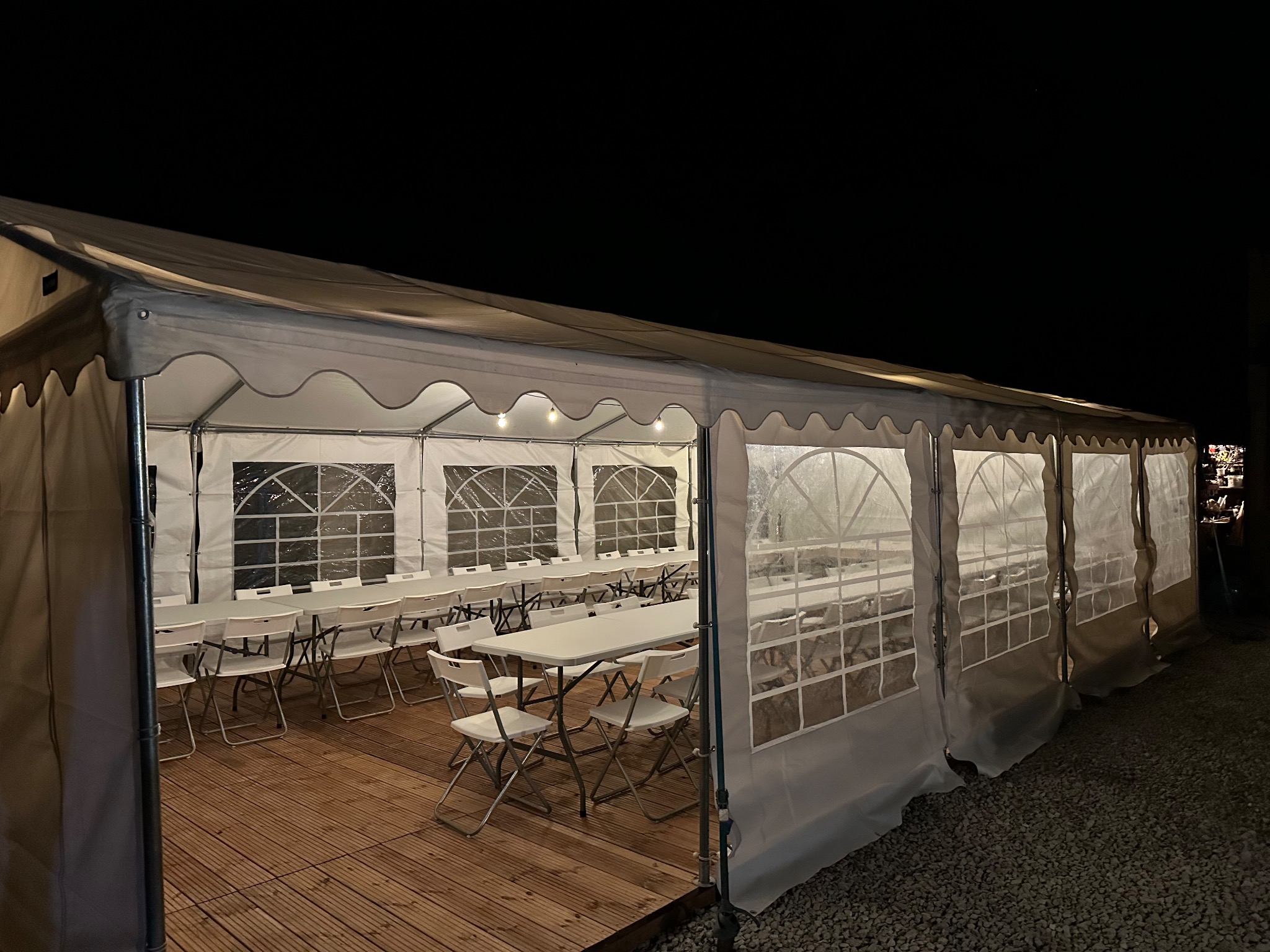 Oświetlony namiot eventowy z drewnianą podłogą i białymi krzesłami ustawionymi wokół stołów, widok nocą z zewnątrz.