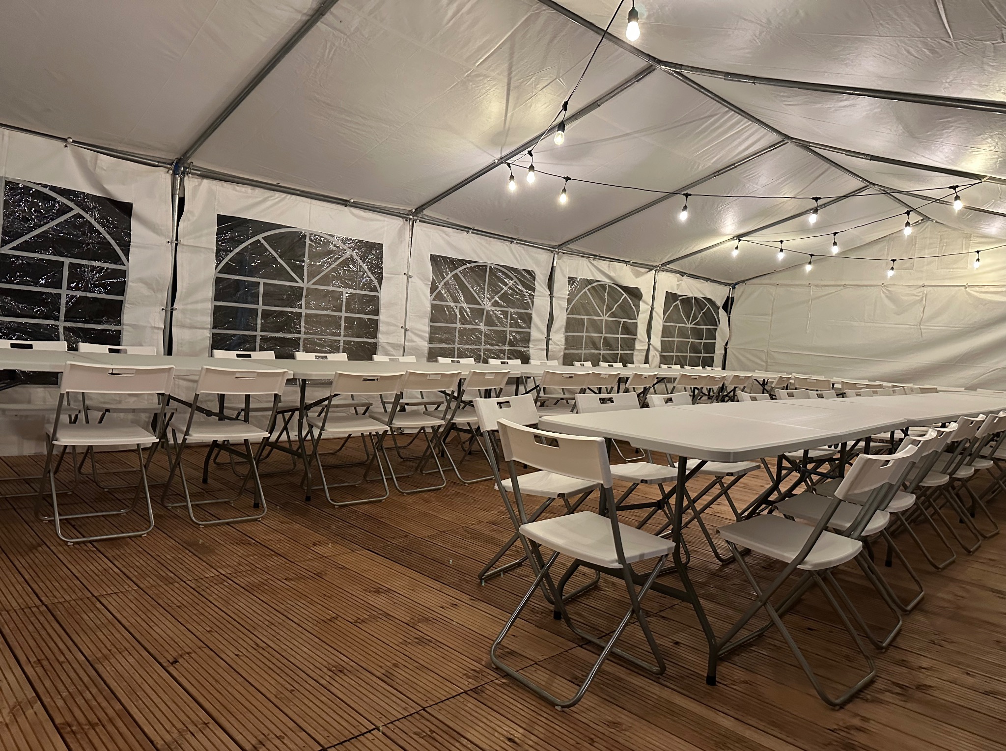 Wnętrze białego namiotu eventowego z drewnianą podłogą, rzędami białych stołów i krzeseł. Dekoracyjne oświetlenie w postaci girlandy żarówek.