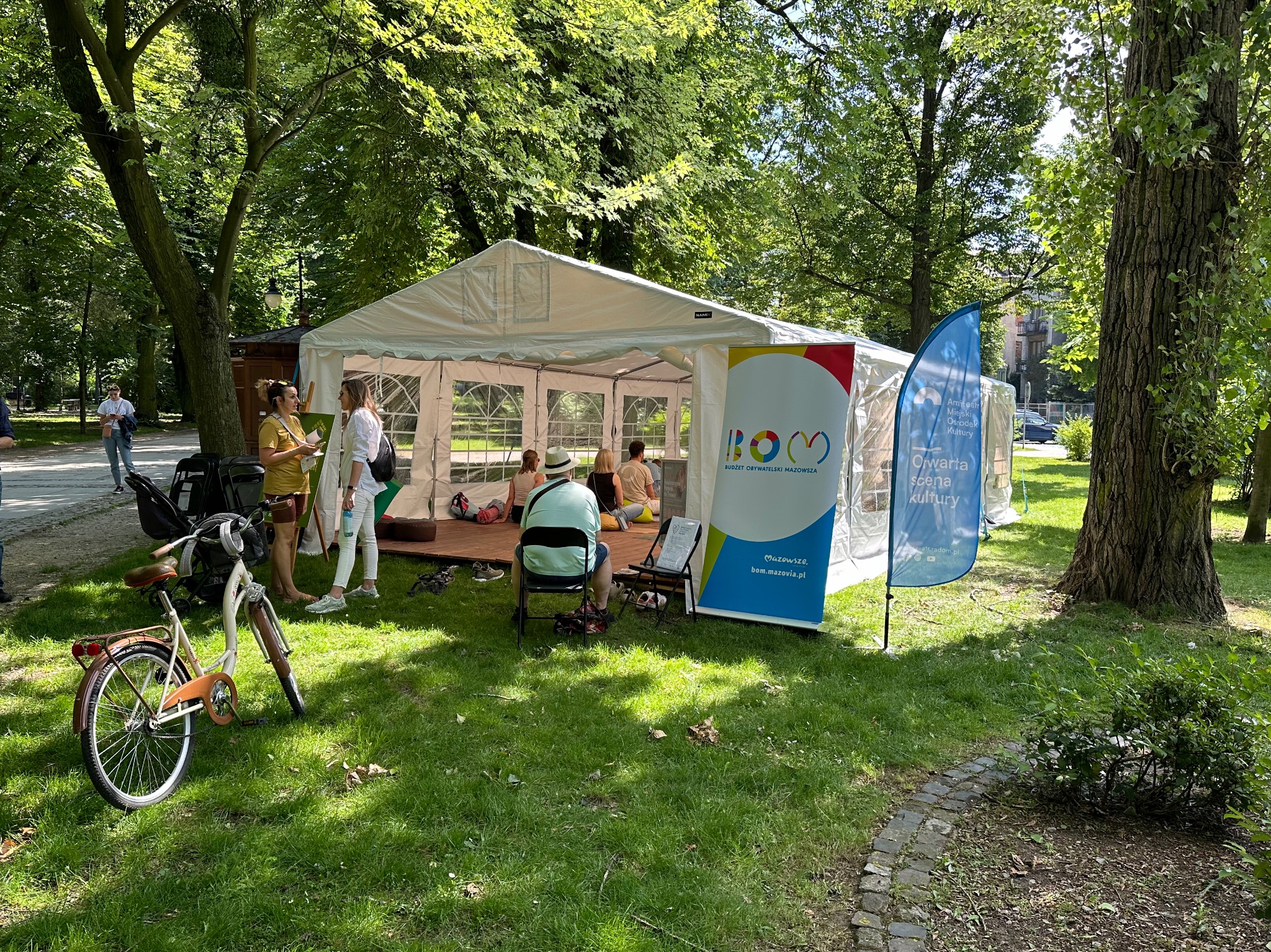 Biały namiot eventowy w parku, z osobami w środku. Obok rower, flagi i tablice informacyjne. Słoneczny dzień, zielona trawa i drzewa w tle.