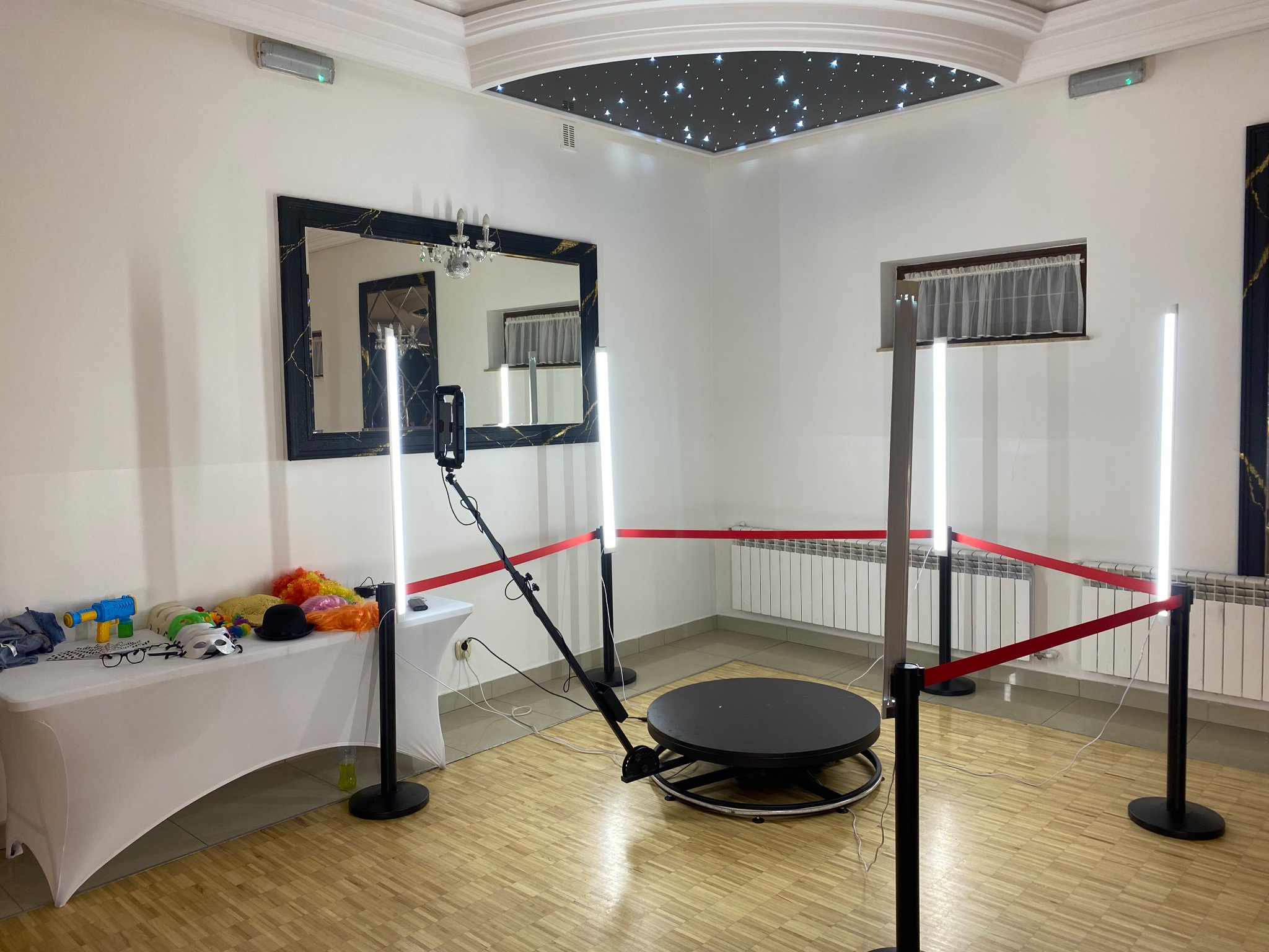 Rotacyjna fotobudka 360 z rekwizytami na stole, otoczona barierkami z czerwonej taśmy w jasnym pomieszczeniu z dekoracyjnym sufitem.
