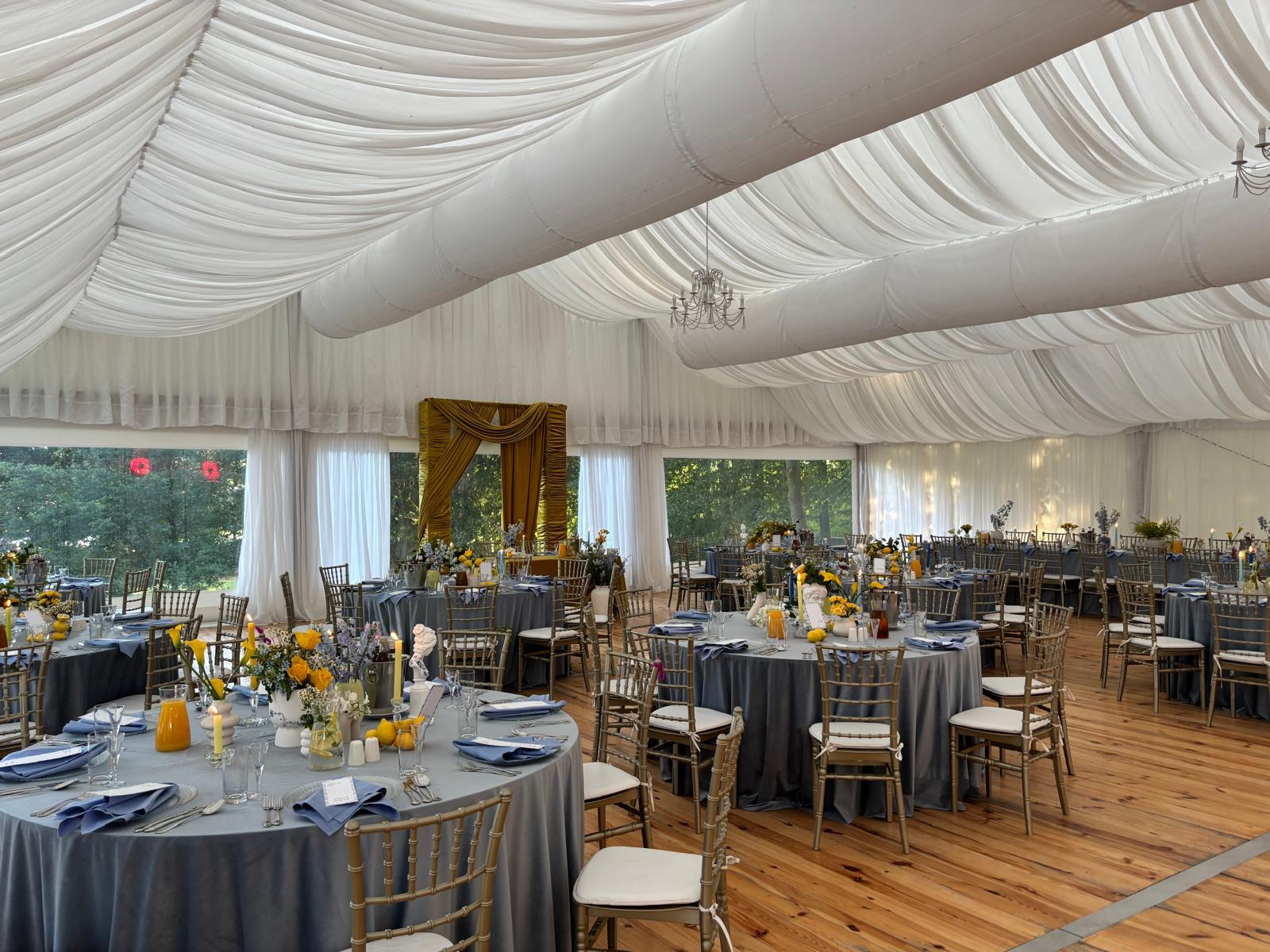 Uroczyste przyjęcie w namiocie: okrągłe stoły z szarymi obrusami, złote krzesła, kwiatowe dekoracje i elegancka zastawa. Biały sufit z draperiami i żyrandol.