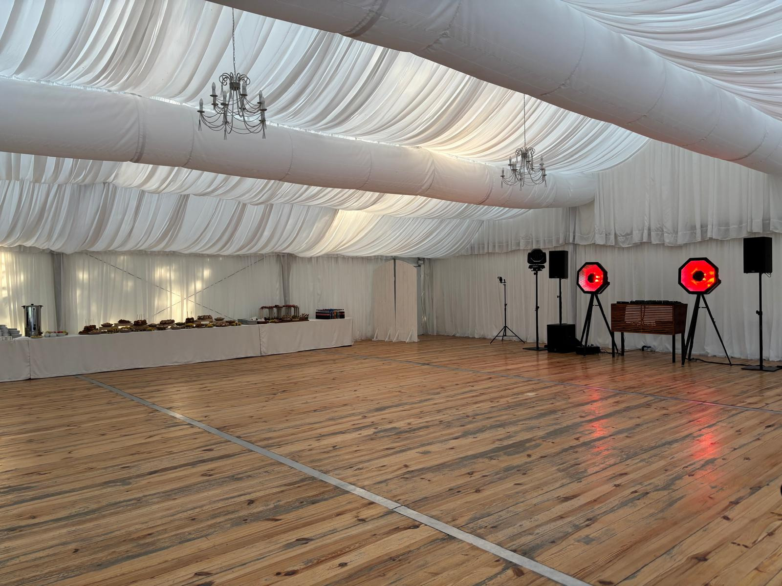 Elegancki namiot bankietowy z drewnianą podłogą, oświetleniem dekoracyjnym i stołem z przekąskami, gotowy na przyjęcie. Widoczne żyrandole i sprzęt nagłośnieniowy.