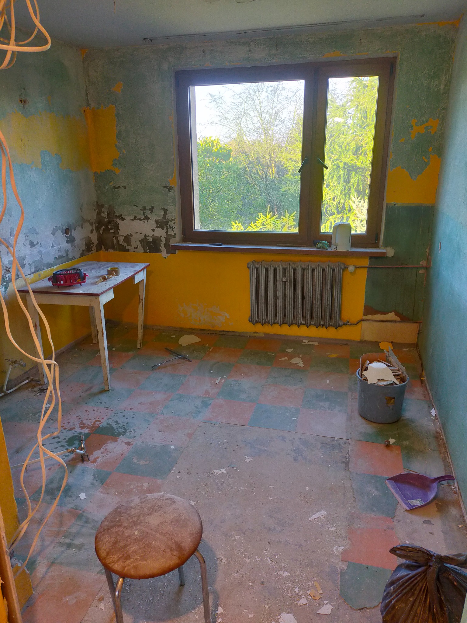 Kadr z remontowanej łazienki: ściany z resztkami farby, okno z widokiem na zieleń, grzejnik, narzędzia. Stan 'przed' z widocznym bałaganem.