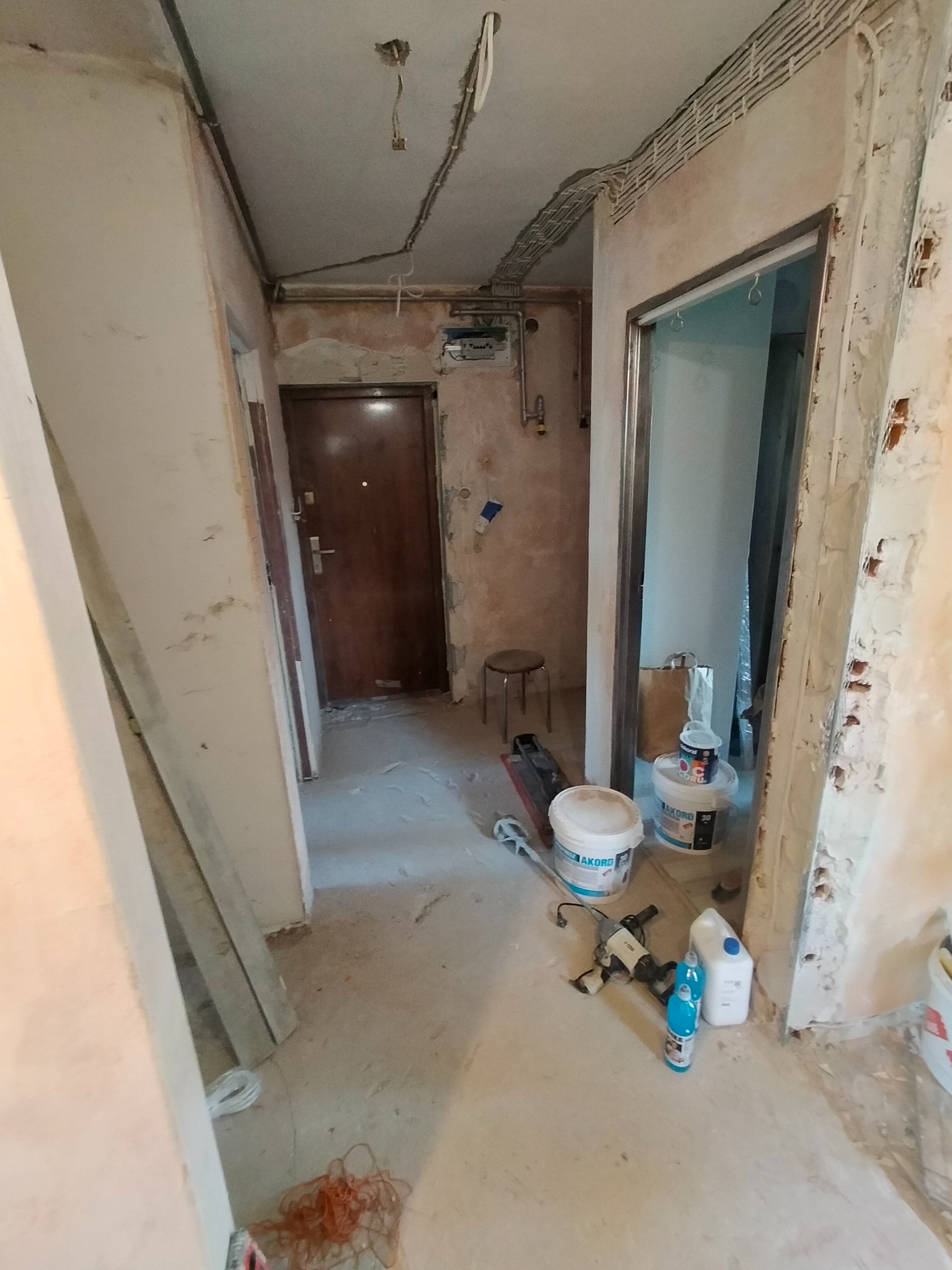 Korytarz w mieszkaniu w trakcie remontu. Widoczne ściany ze śladami starej farby, odkryte przewody elektryczne, narzędzia i materiały budowlane.