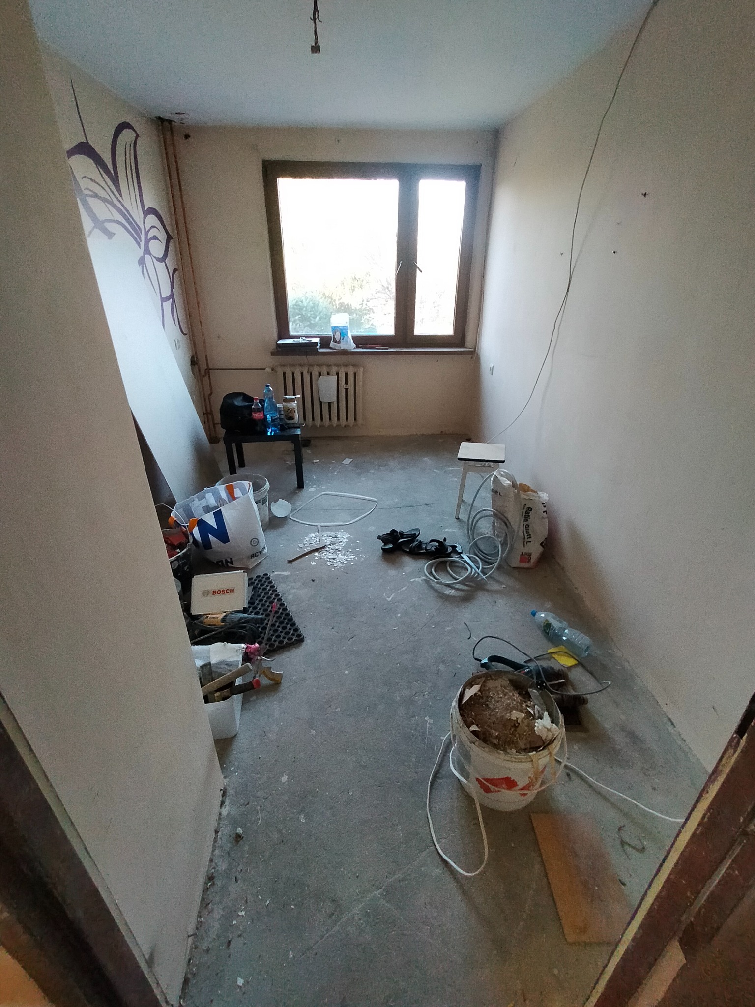 Pokój w trakcie remontu: surowe ściany, okno, narzędzia i materiały budowlane. Widoczne ślady po wcześniejszych pracach. Stan 'przed' wykończeniem wnętrza.