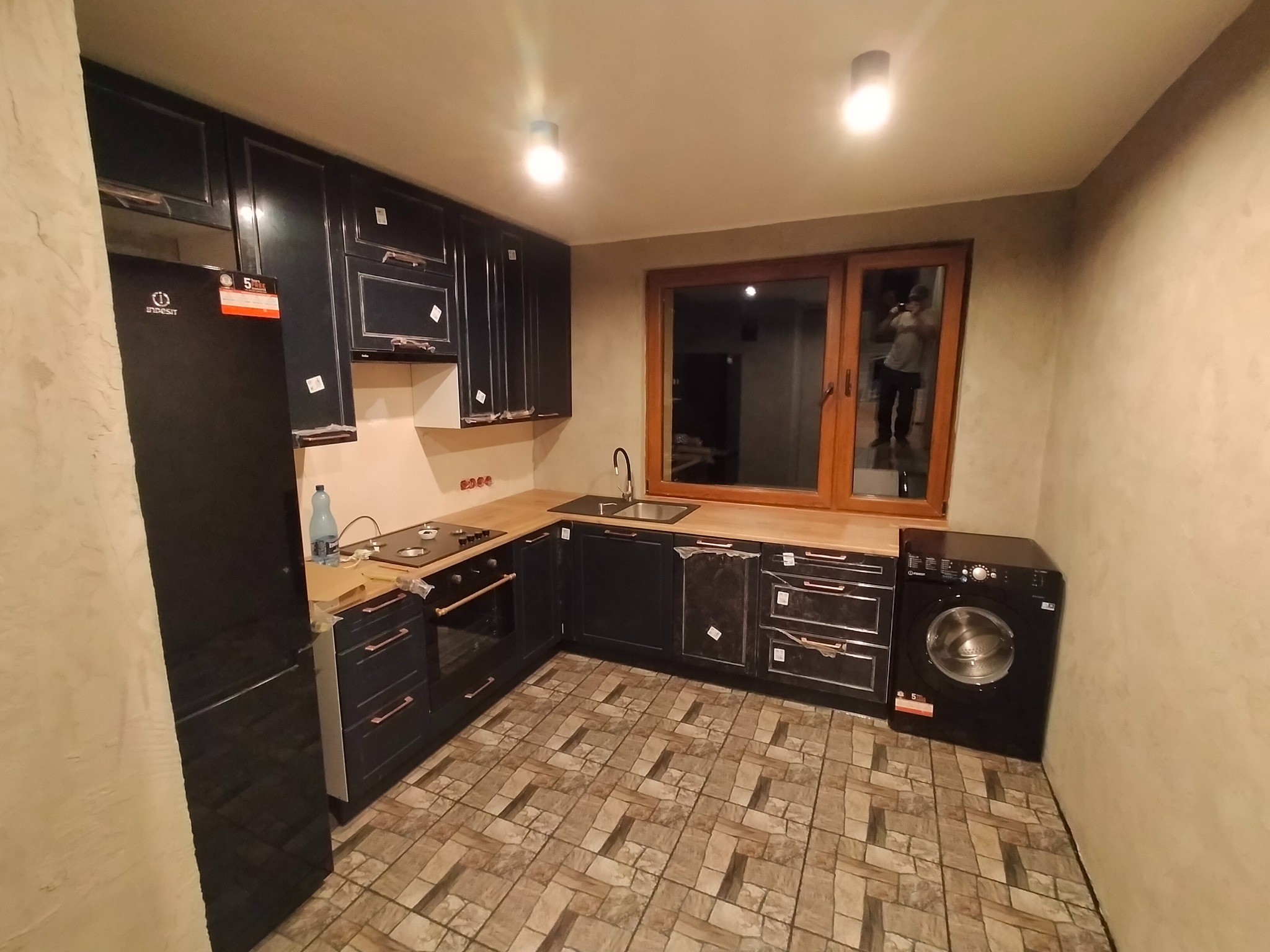 Ciemna zabudowa kuchenna z drewnianym blatem, płyta grzewcza i zlew. Na podłodze wzorzyste płytki. Widoczna pralka i lodówka. Częściowo zaklejone folią ochronną.