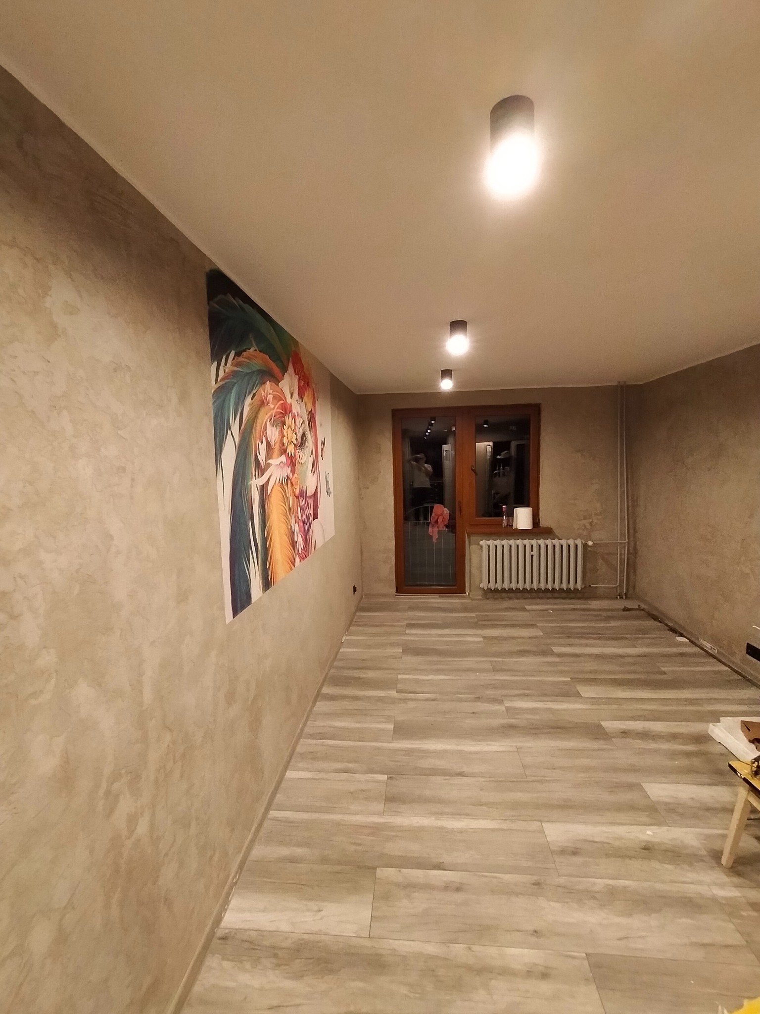 Wykończone wnętrze pokoju z panelami podłogowymi imitującymi drewno i ścianami o fakturze betonu. Na ścianie wisi duży obraz.