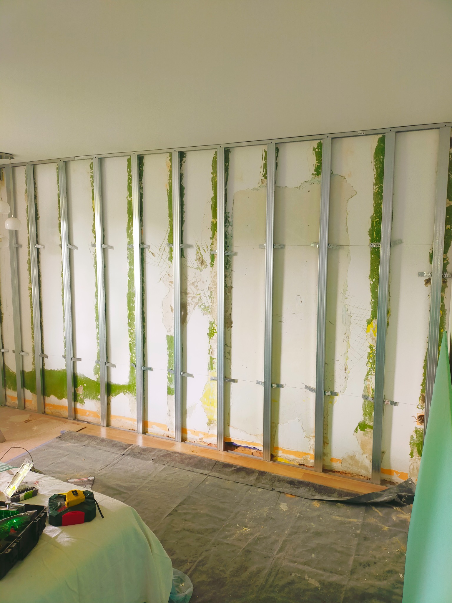 Montaż metalowego stelażu pod płytę karton-gips na starej ścianie z widocznymi śladami farby i tapety. Na podłodze rozłożona folia ochronna i narzędzia.