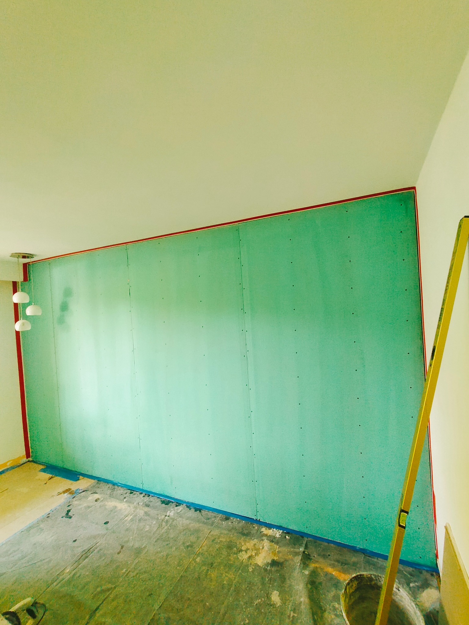 Ściana z zielonych płyt karton-gips, zabezpieczona taśmą, z widoczną poziomicą i lampą sufitową. Etap prac przed wykończeniem.
