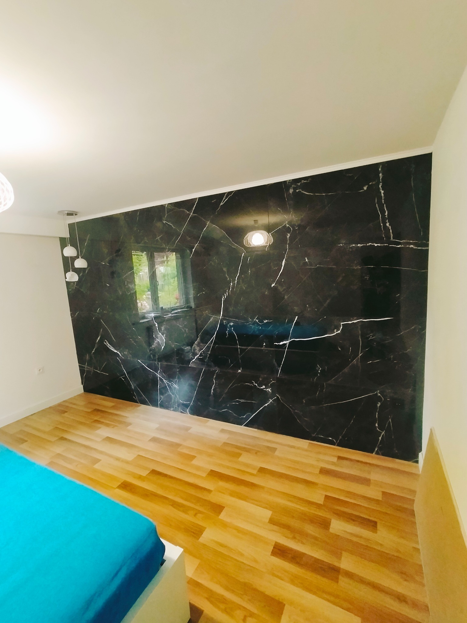 Wykończone wnętrze sypialni z czarną ścianą imitującą marmur i drewnianą podłogą. Okno z widokiem na zieleń. Nowoczesne oświetlenie sufitowe.