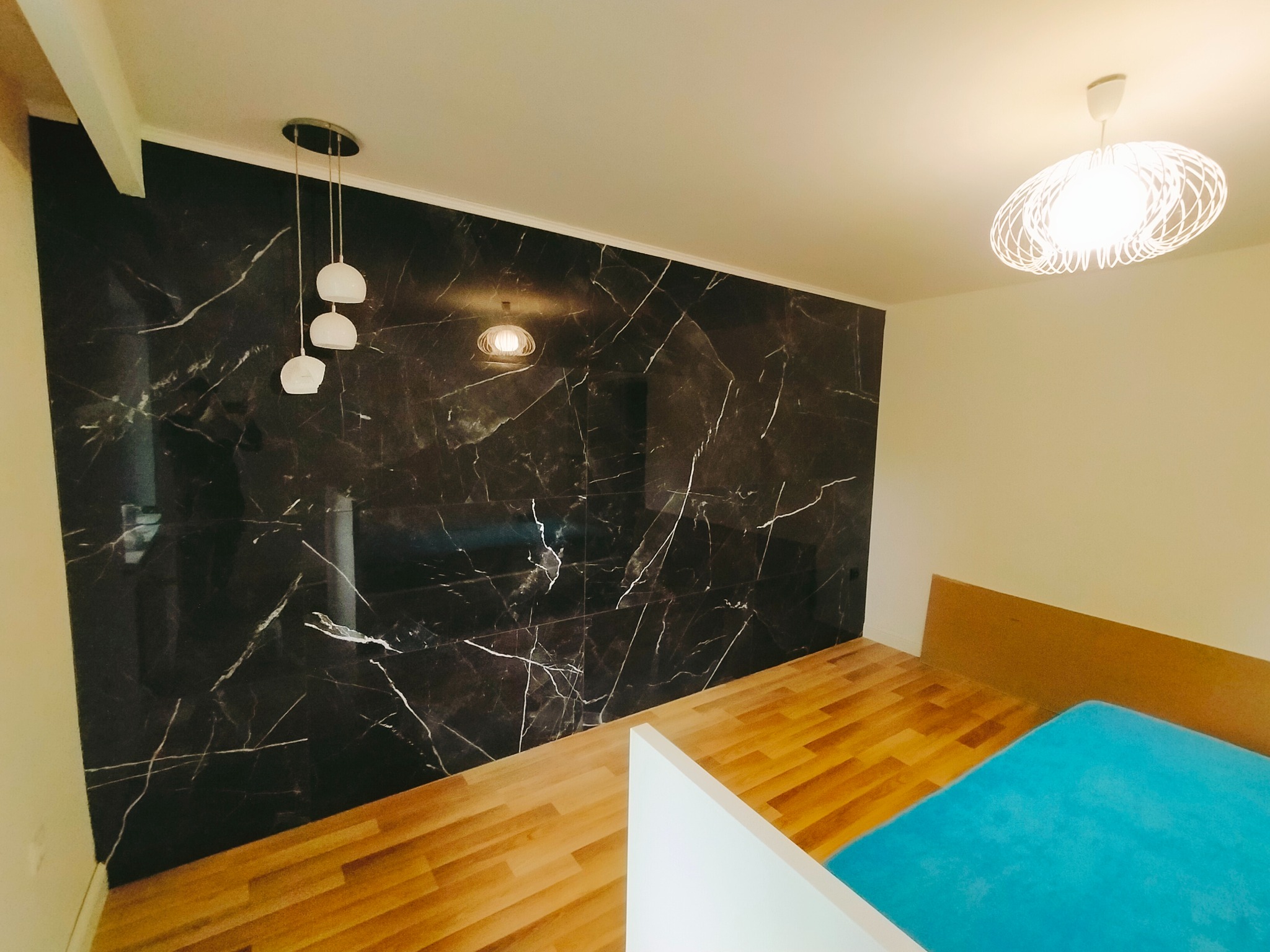 Wykończony pokój z panelami podłogowymi i ścianą z płytek imitujących czarny marmur. Widoczne lampy sufitowe.