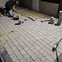 Mkb-Bruk - Prace brukarskie w toku: ułożona część szarej kostki brukowej, stosy kostki przygotowane do ułożenia, widoczny pracownik w trakcie układania, piasek pod kostką, szczotka brukarska w prawym dolnym...