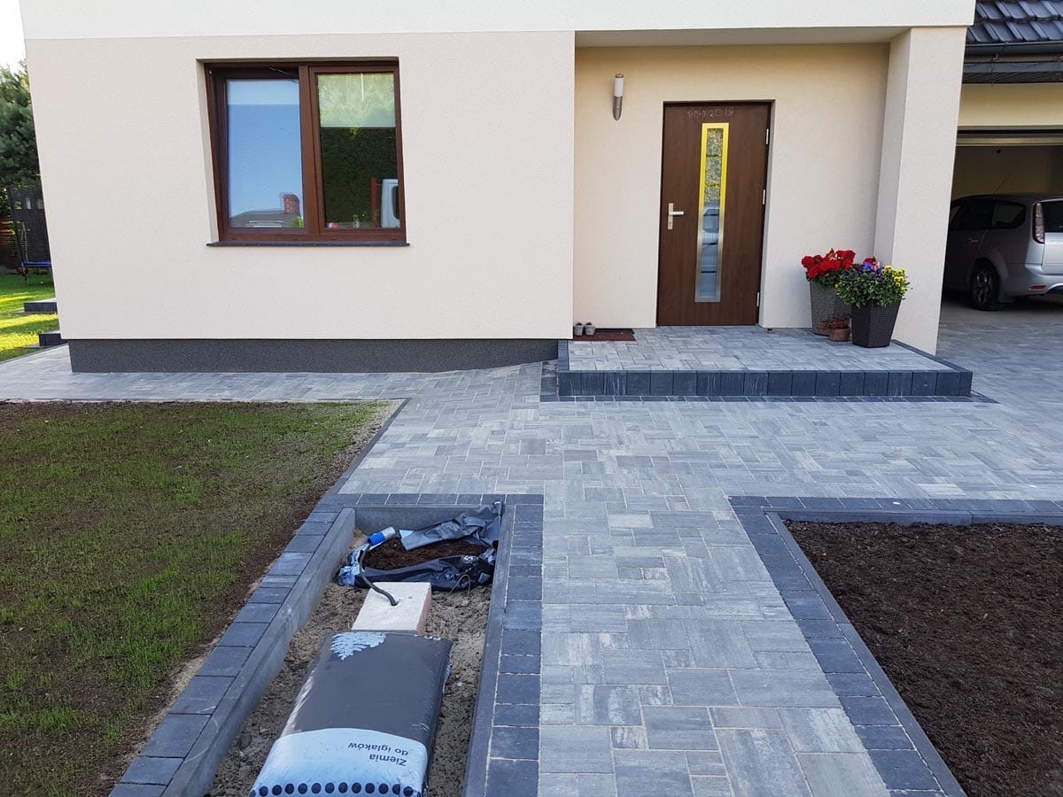 Nowoczesny podjazd z szarej kostki brukowej ułożonej we wzór jodełki, obrzeża z ciemnej kostki, widoczny fragment trawnika i rabaty z ziemią, dom jednorodzinny w tle.