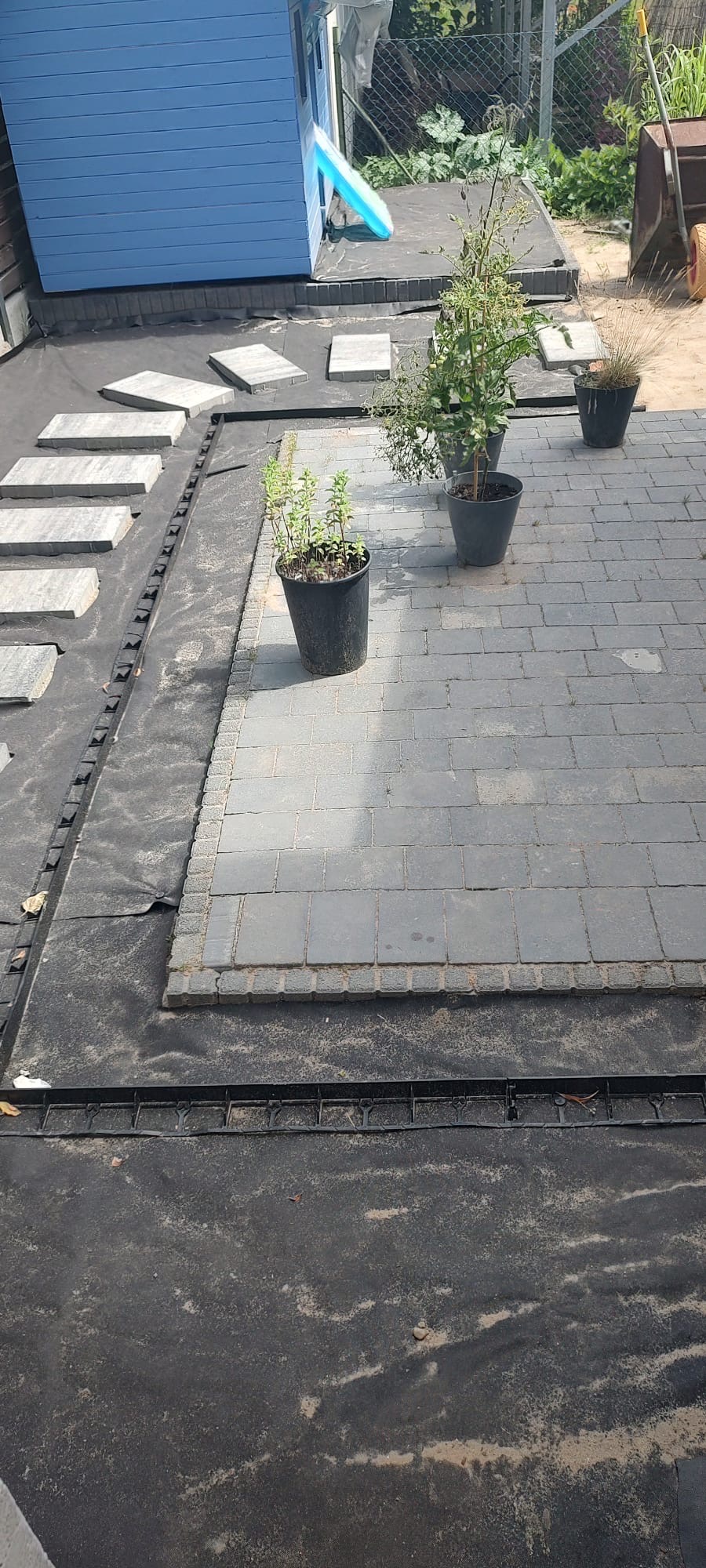 Fragment układanej nawierzchni z szarej kostki, widoczne czarne geowłókniny i plastikowe obrzeża, w tle mały, niebieski domek dla dzieci i donice z roślinami.