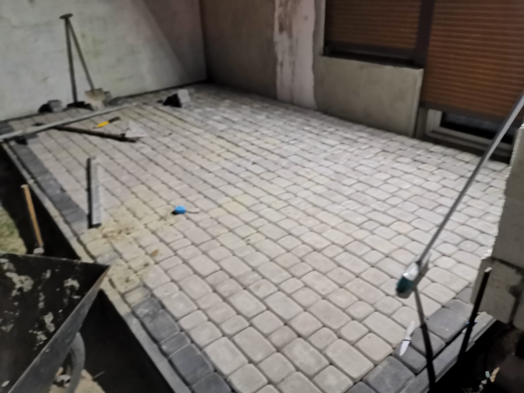 Fragment układanej powierzchni z jasnej kostki brukowej z ciemnoszarą obwódką, widoczne narzędzia pracy i taczka w rogu kadru.