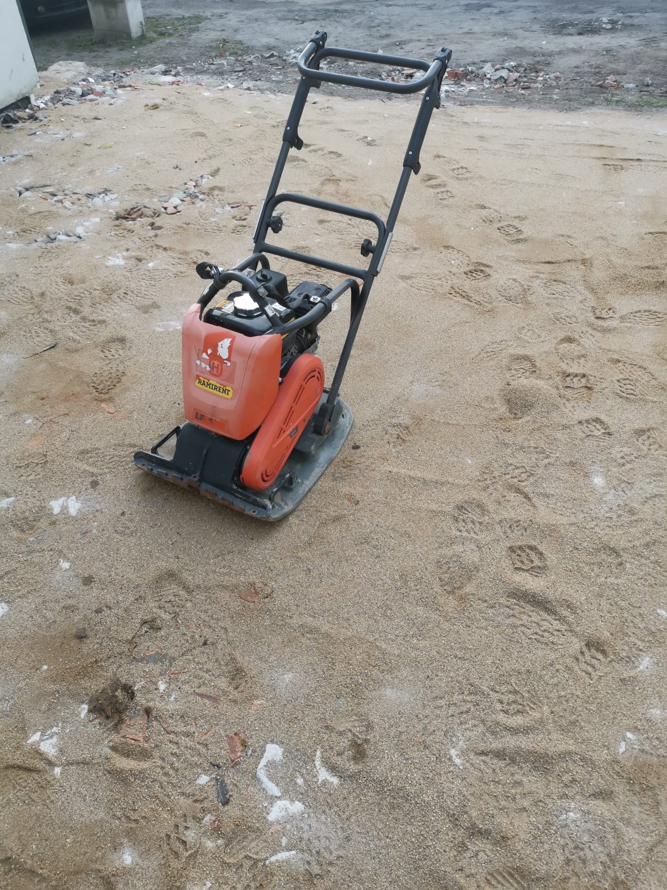 Pomarańczowa ubijarka spalinowa marki Ramirent na piasku, widoczne ślady stóp i gruz.