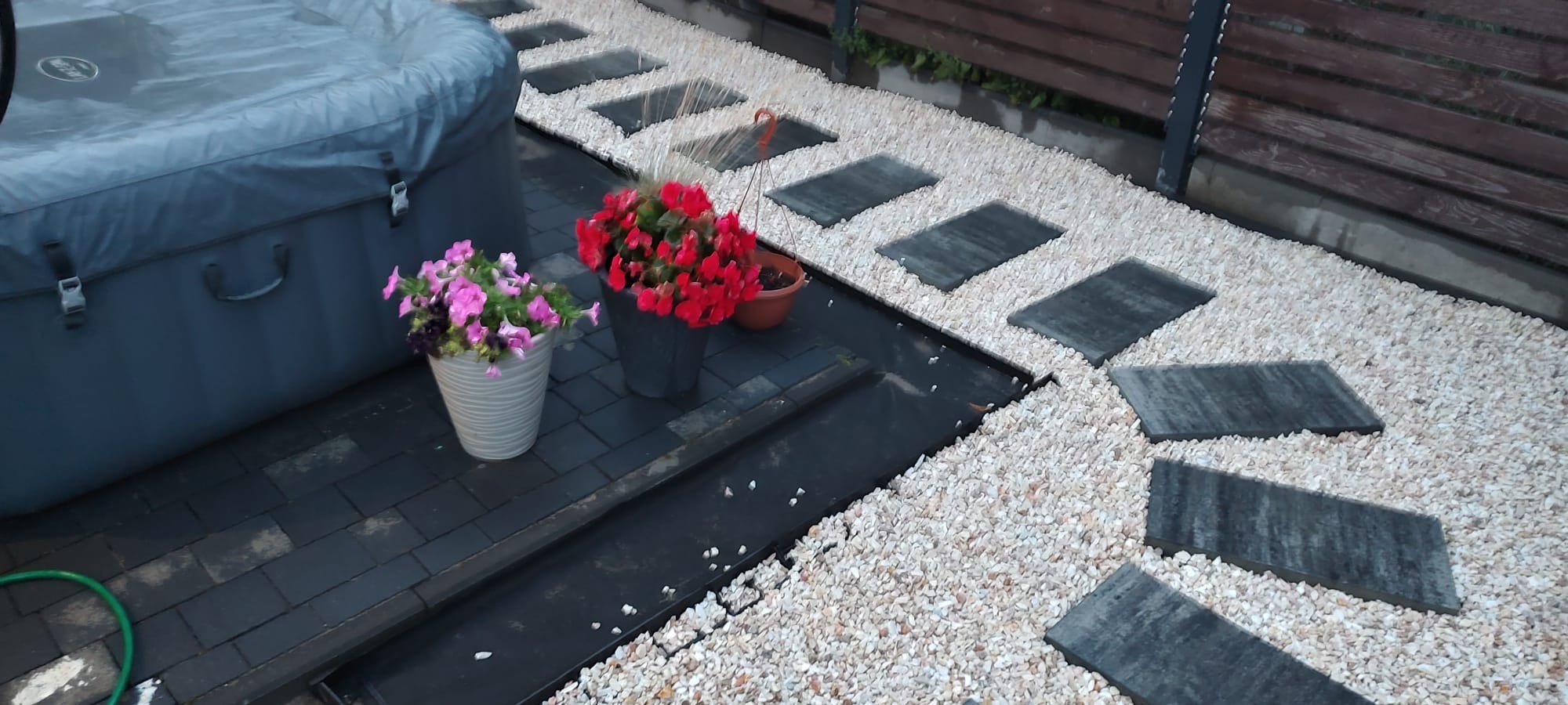 Aranżacja przestrzeni przydomowej z ścieżką z prostokątnych płyt i białego grysu, obok jacuzzi i donic z kwiatami.