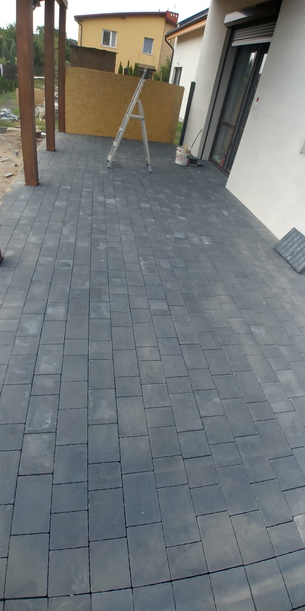 Ułożona szara kostka brukowa na tarasie, z widoczną drabiną i elementami konstrukcji z drewna w tle, częściowo ukończona realizacja.