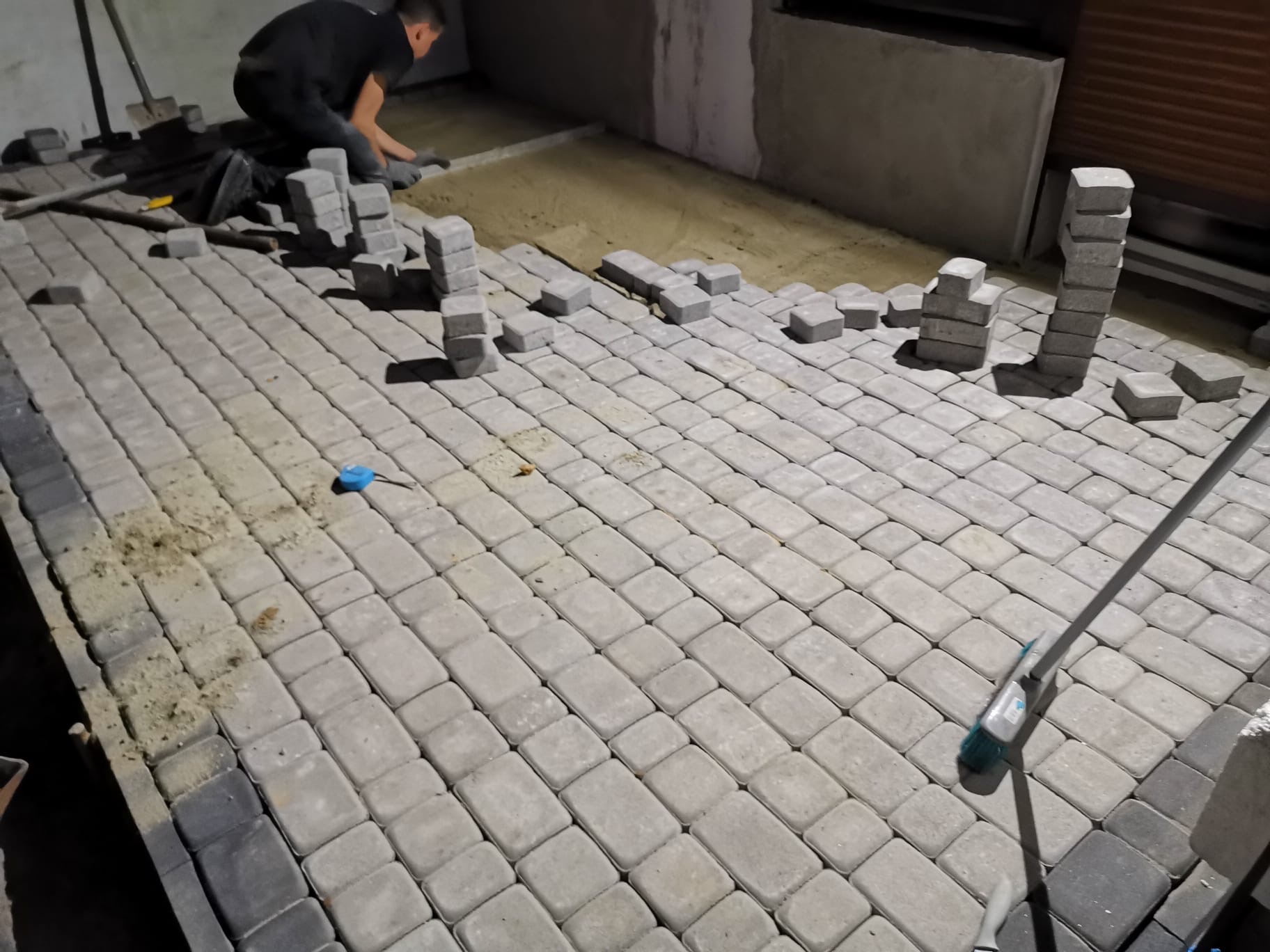 Prace brukarskie w toku: ułożona część szarej kostki brukowej, stosy kostki przygotowane do ułożenia, widoczny pracownik w trakcie układania, piasek pod kostką, szczotka brukarska w prawym dolnym...