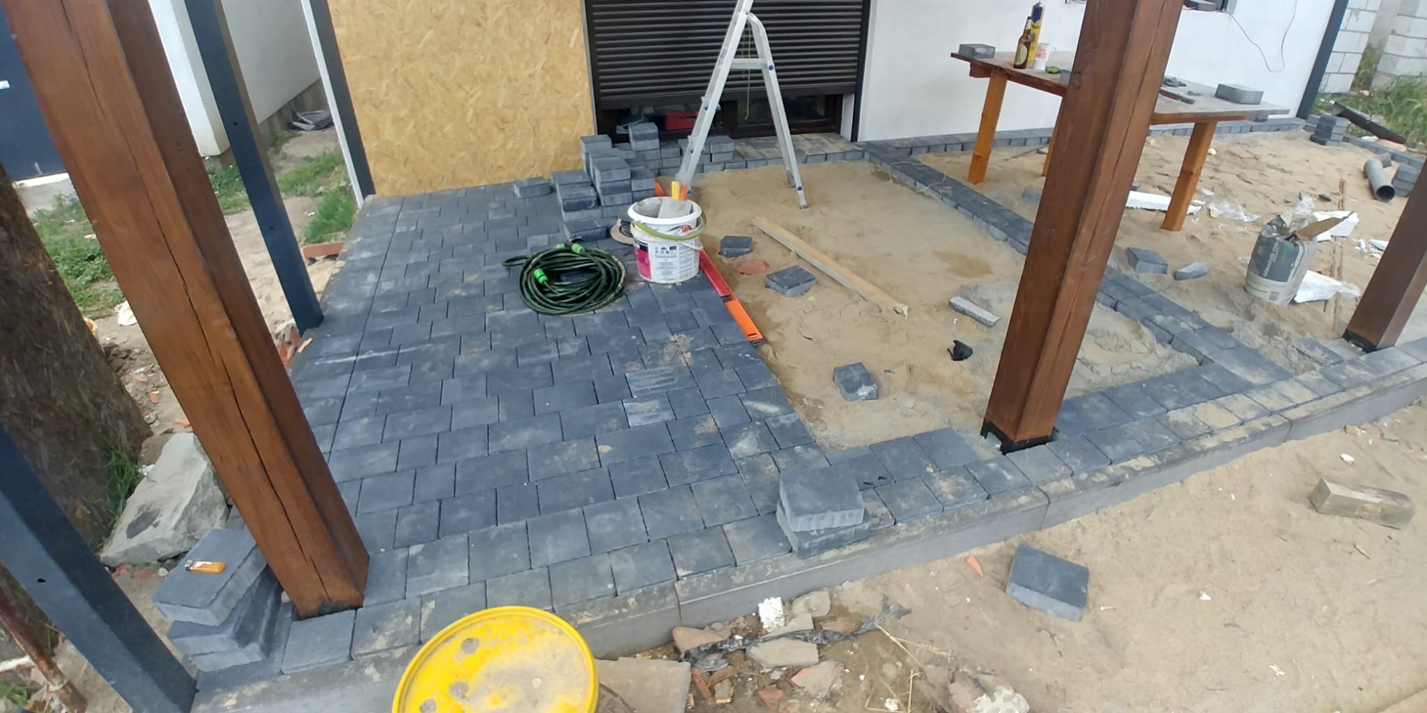 Etap układania ciemnoszarej kostki brukowej wokół drewnianych słupów konstrukcji z widocznym wiadrem z farbą, wężem ogrodowym i poziomnicą.