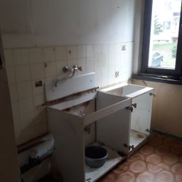 Marek Kowalski - Demontaż szafek kuchennych w trakcie remontu: widoczne stare płytki, odsłonięte rury i przygotowane do wymiany podłoże. Przez okno widoczny budynek mieszkalny i samochód.