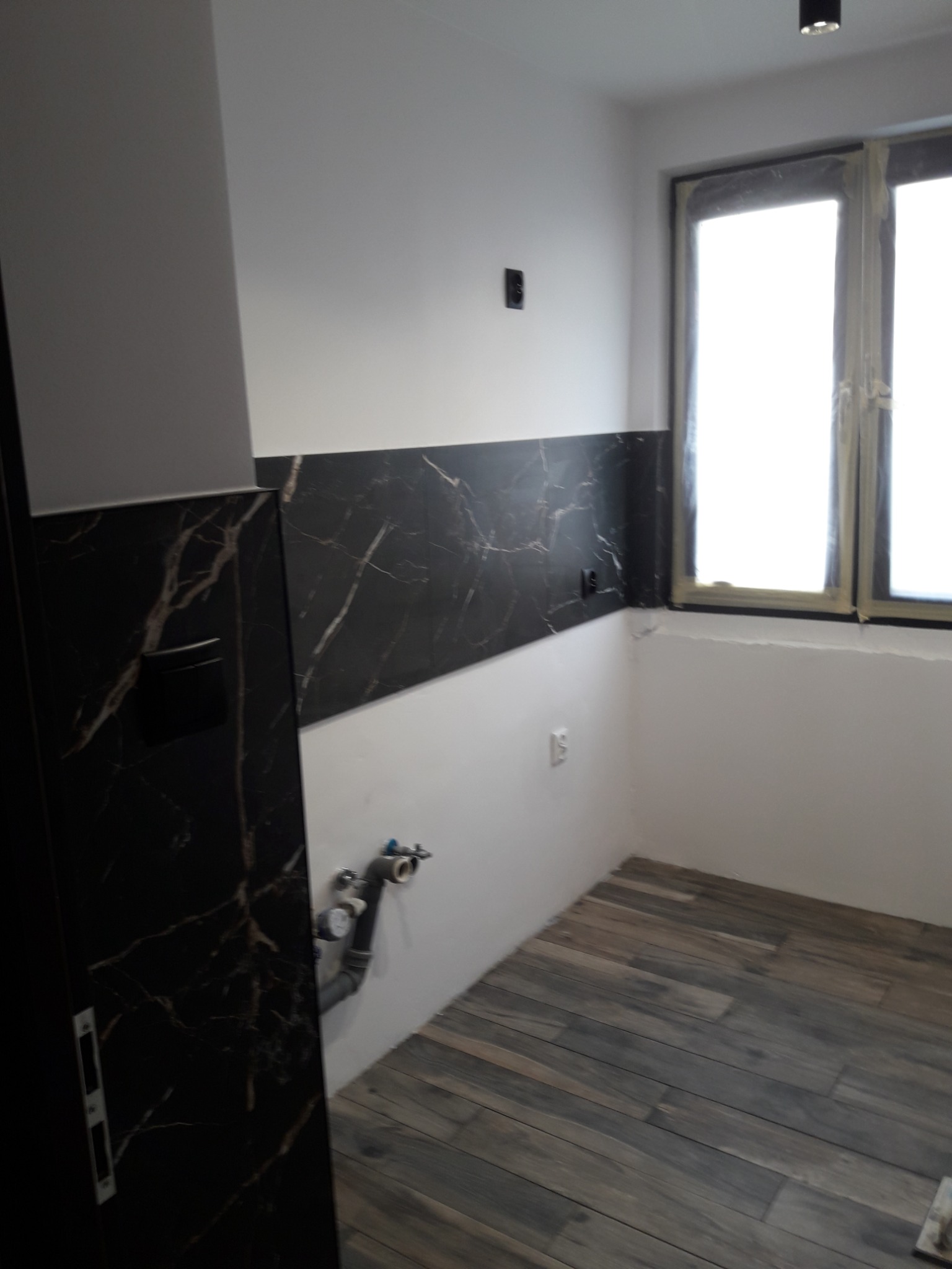 Łazienka w trakcie remontu: położona podłoga z płytek imitujących drewno, ściany częściowo wyłożone czarnymi płytkami z białym marmurkowym wzorem, widoczne rury instalacji hydraulicznej i okno...