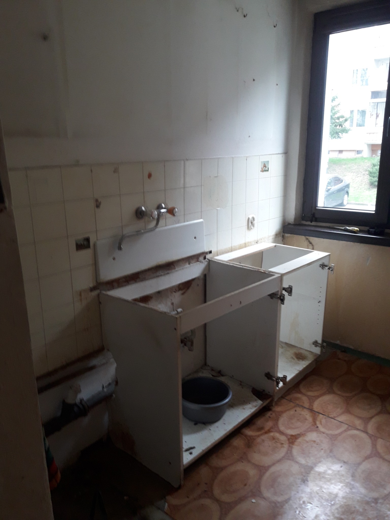 Demontaż szafek kuchennych w trakcie remontu: widoczne stare płytki, odsłonięte rury i przygotowane do wymiany podłoże. Przez okno widoczny budynek mieszkalny i samochód.