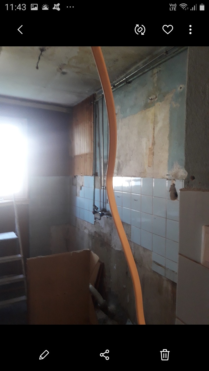 Łazienka w trakcie remontu: zerwane płytki, widoczne instalacje hydrauliczne, stara armatura, pomarańczowy wąż i kartonowe pudełko na podłodze.