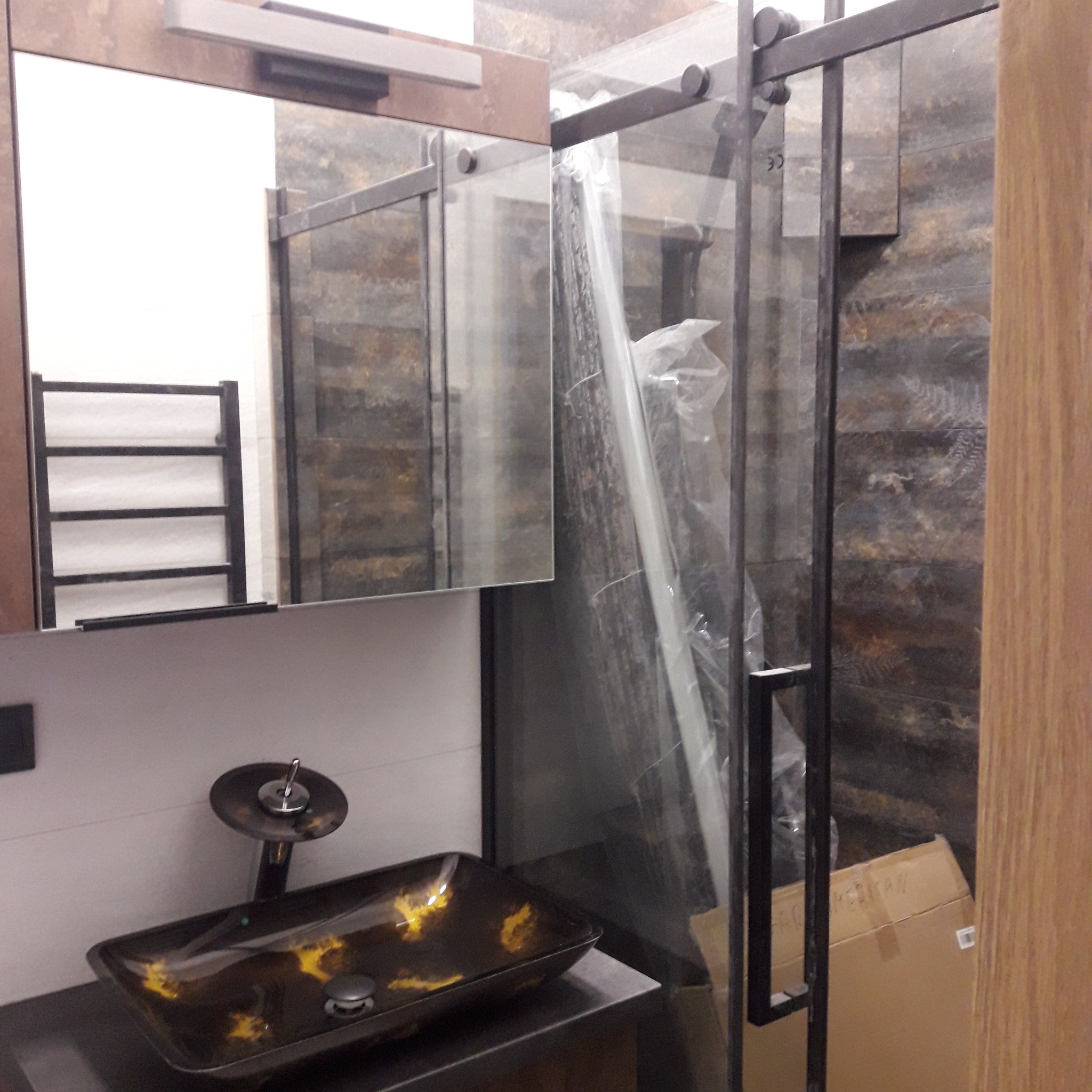 Nowoczesna łazienka w trakcie wykańczania: szklana kabina prysznicowa z czarnymi ramami, umywalka z brązowego szkła z złotymi akcentami, lustro z oświetleniem i grzejnik drabinkowy widoczny...