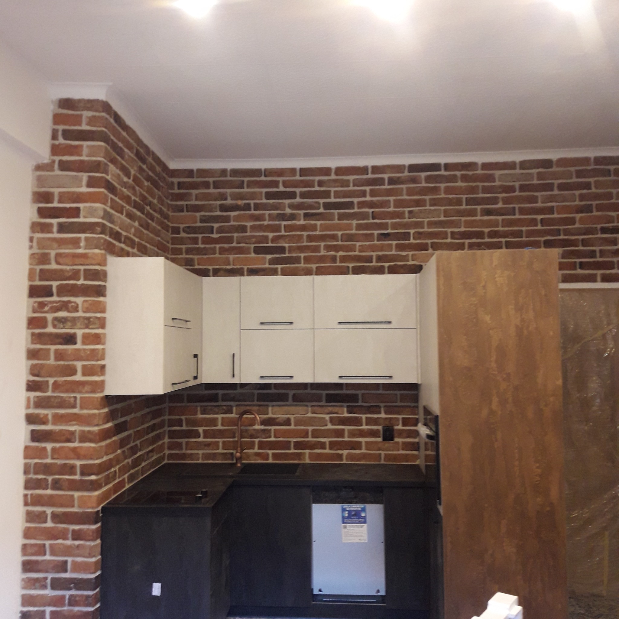 Kuchnia z ceglaną ścianą, białymi szafkami wiszącymi z czarnymi uchwytami, ciemnym blatem i miedzianą baterią.