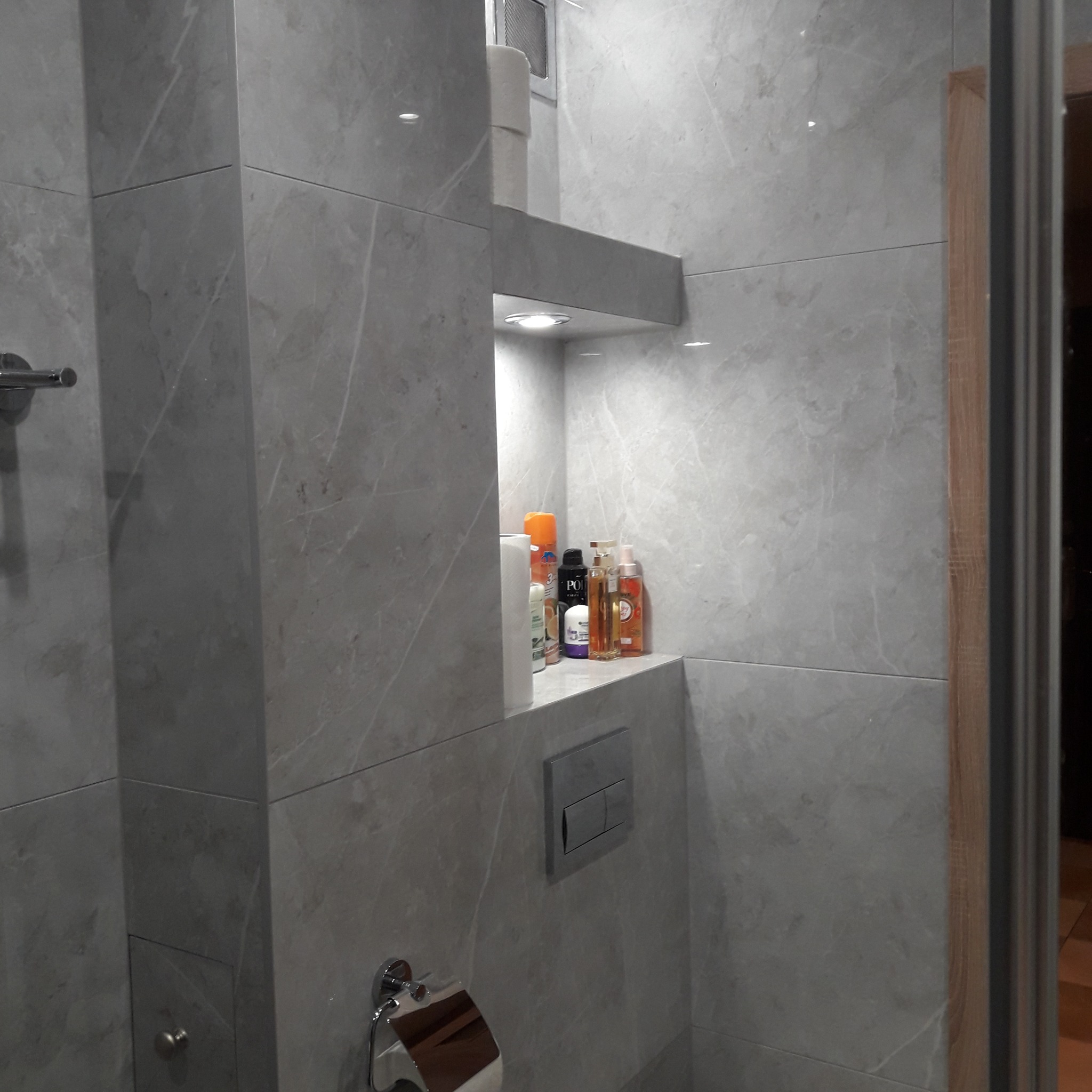 Wykończona łazienka z szarymi płytkami imitującymi marmur, wnęka z oświetleniem i kosmetykami, chromowany uchwyt na papier toaletowy oraz przycisk spłuczki.