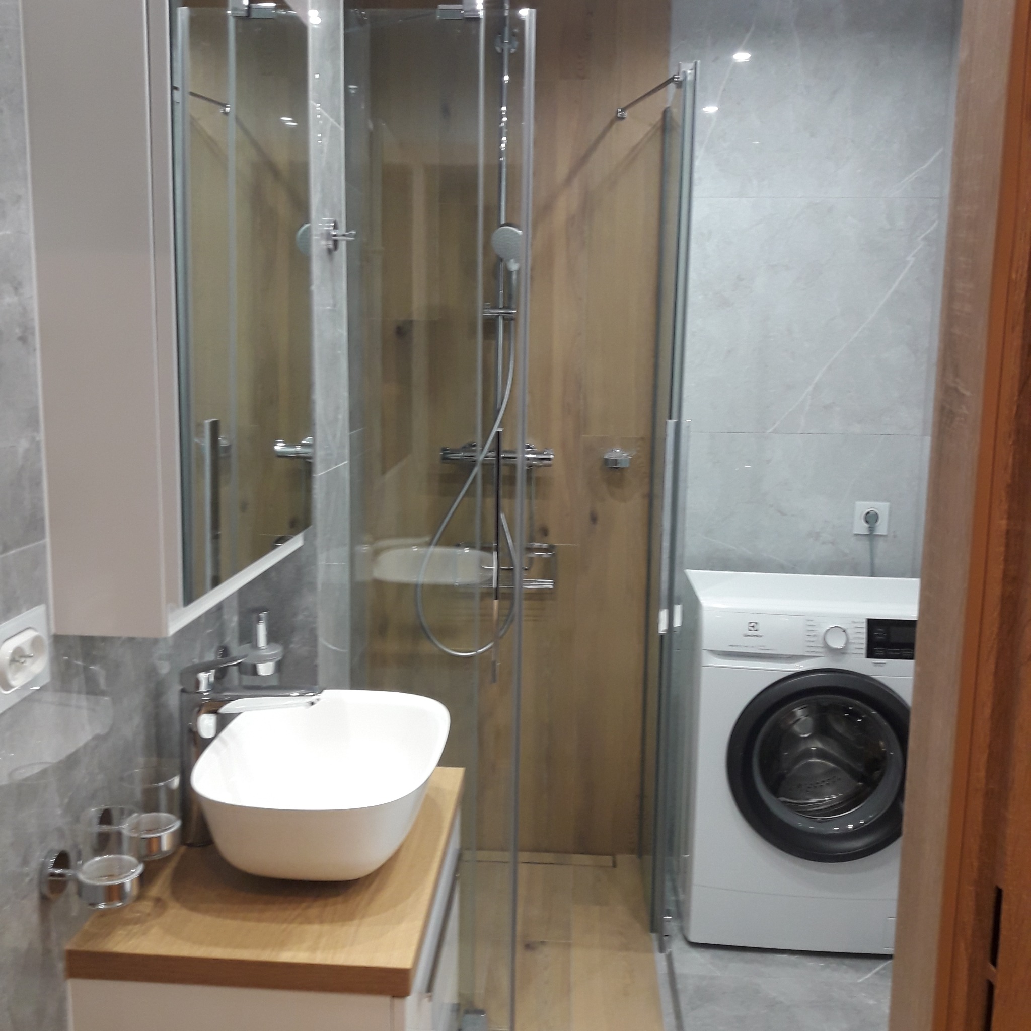 Nowoczesna łazienka z kabiną prysznicową ze szklanymi drzwiami, umywalką nablatową na drewnianym blacie, pralką Electrolux i szarą marmurową okładziną ścian.