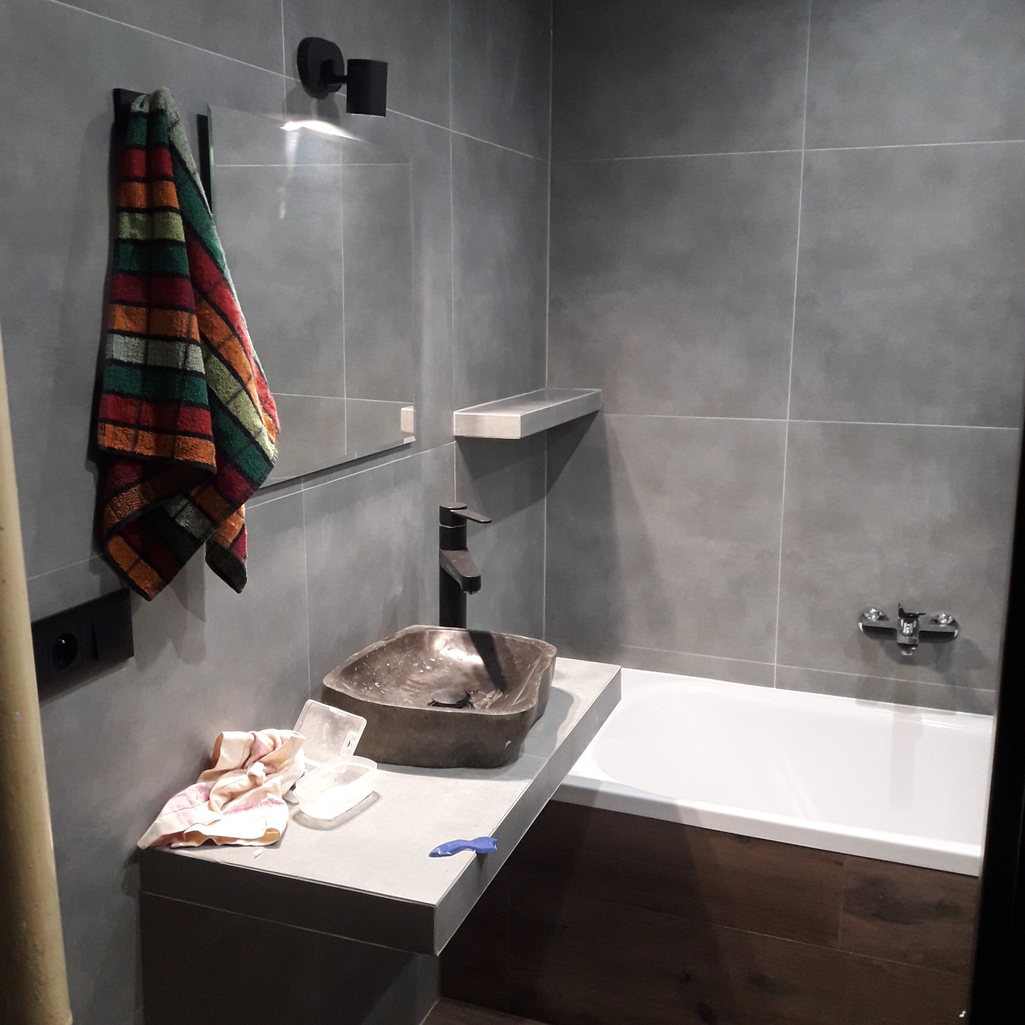 Umywalka z kamienia w szarej łazience w Krakowie, z czarną baterią i ręcznikiem w kratę, widoczna wanna i akcesoria glazurnicze.