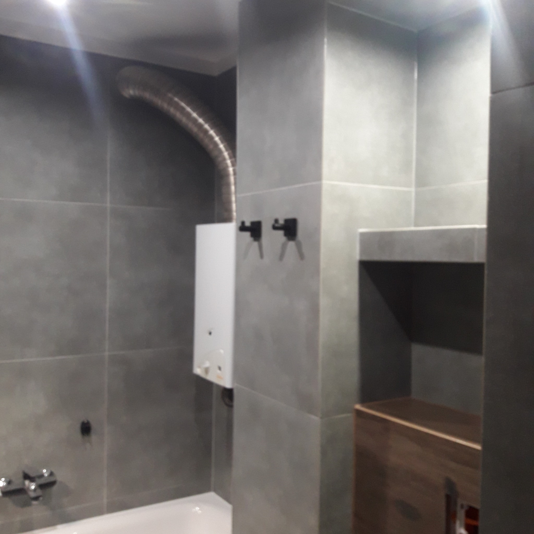 Szara łazienka z zamontowanym bojlerem podłączonym do komina wentylacyjnego, widoczne płytki ceramiczne, bateria umywalkowa oraz szafka z półką.