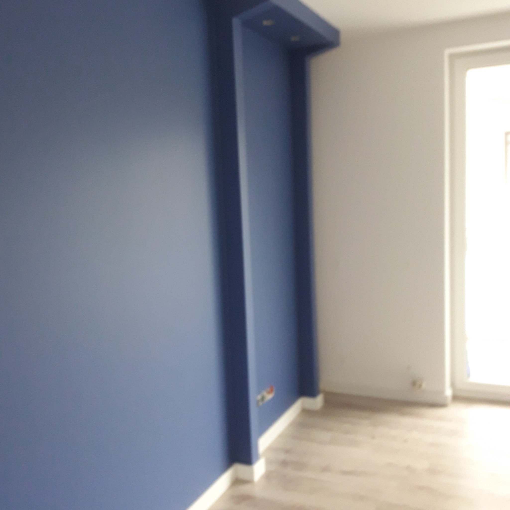 Ściana pomalowana na niebiesko z ozdobnym elementem architektonicznym i gniazdkiem elektrycznym, widoczne fragmenty podłogi i okna.