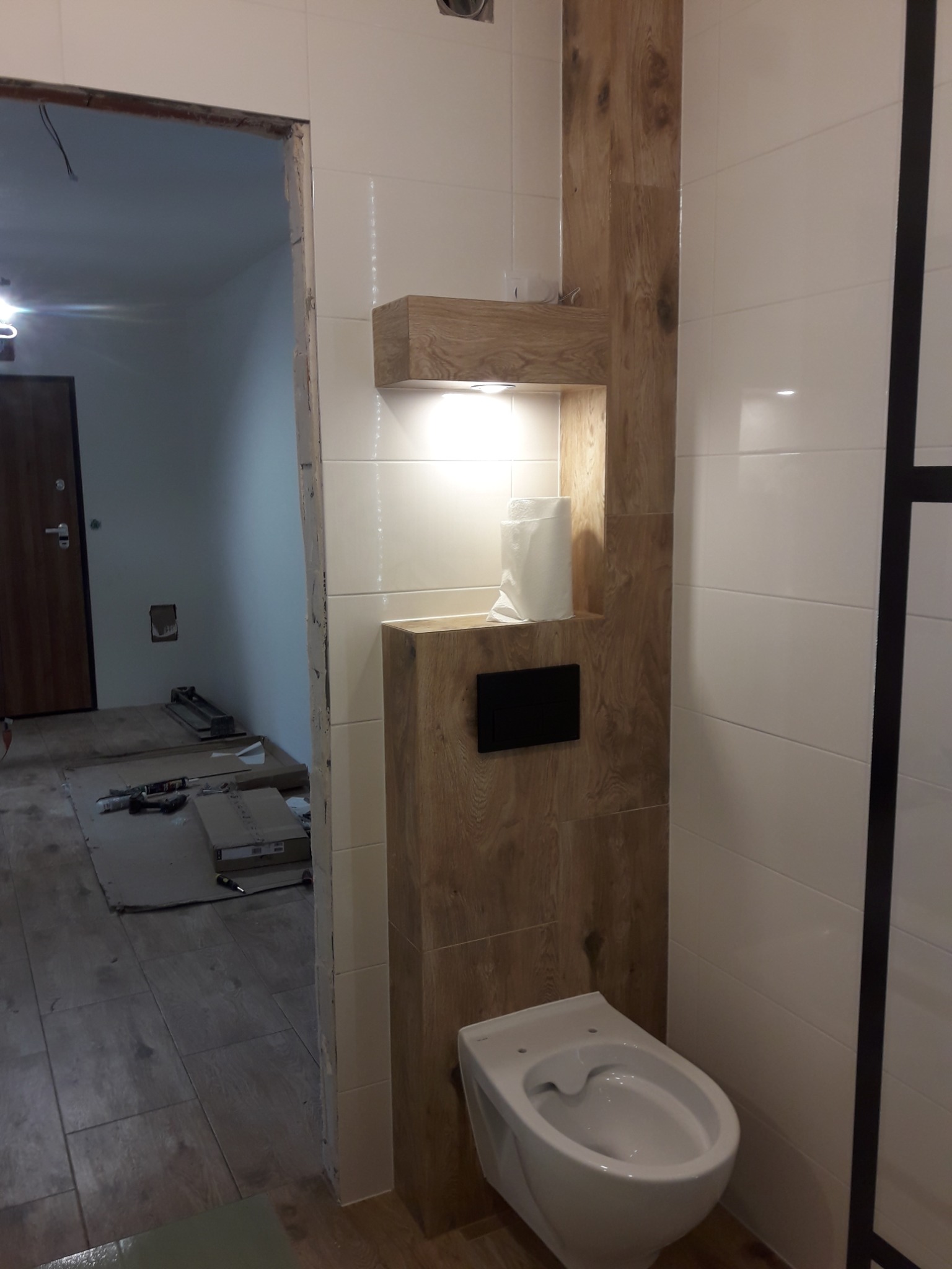 Wnętrze łazienki w trakcie wykańczania, widoczna podwieszana toaleta, zabudowa z drewnopodobnych paneli z półką podświetlaną LED, białe płytki na ścianach oraz narzędzia i materiały budowlane w tle.