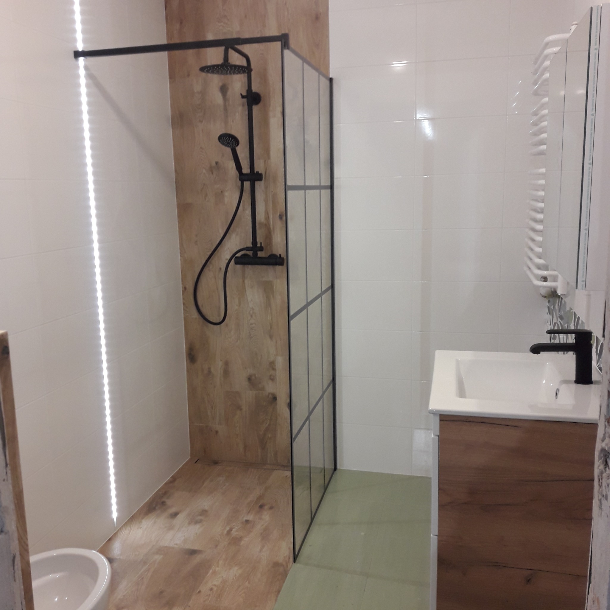 Nowoczesna łazienka z kabiną prysznicową typu walk-in, czarną armaturą i białymi płytkami, akcent drewnopodobny na ścianie i podłodze w strefie prysznica.