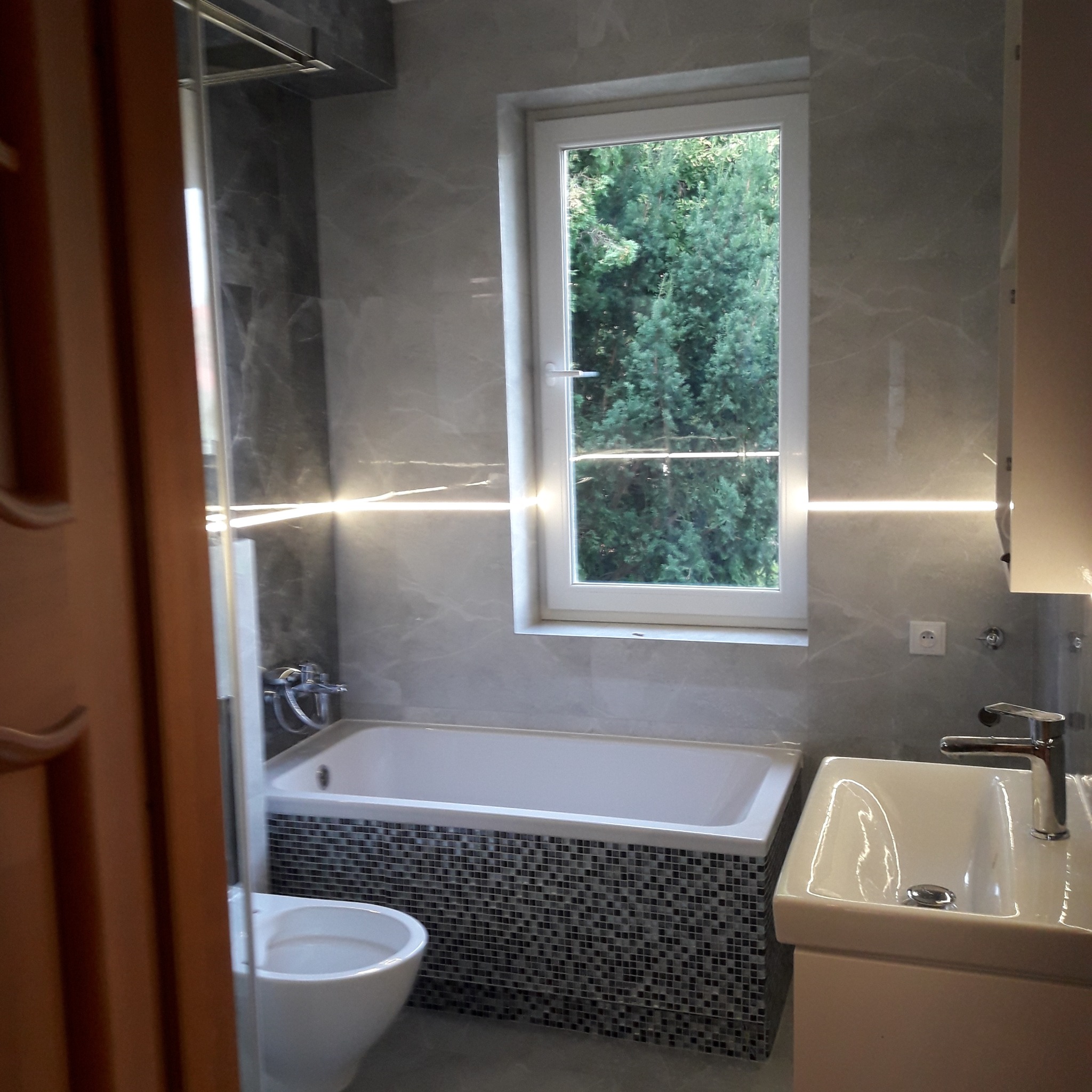 Nowoczesna łazienka z wanną obłożoną mozaiką, umywalką nablatową i oknem z widokiem na zieleń, oświetlona listwami LED.