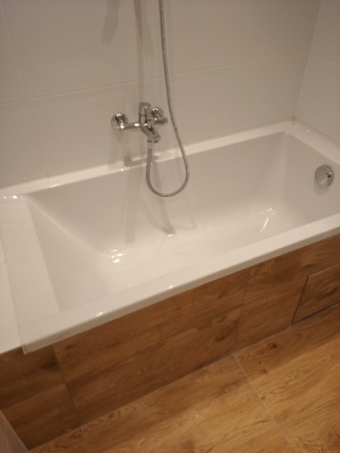 Biała, prostokątna wanna z chromowaną baterią i słuchawką prysznicową, obudowana panelami imitującymi drewno, na tle białych płytek i podłogi z paneli drewnopodobnych.