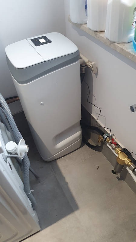 Nowoczesny zmiękczacz wody z elektronicznym panelem sterowania, podłączony do instalacji wodnej z filtrem i zaworem kulowym, umieszczony w pomieszczeniu gospodarczym obok pralki.