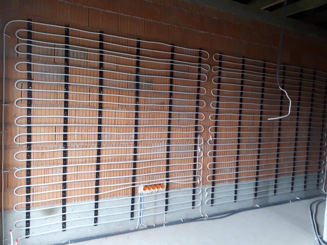 Rury ogrzewania podłogowego zamocowane do ściany z pustaków, widoczna skrzynka elektryczna i przewody.