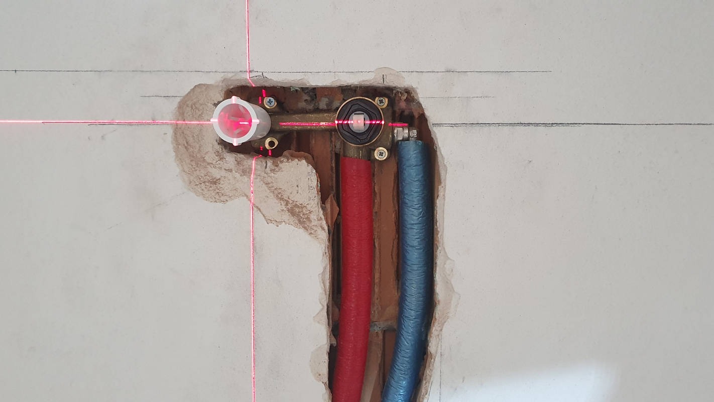 Wnęka w ścianie z zamontowaną baterią podtynkową, widoczne rury doprowadzające wodę oraz czerwona wiązka lasera poziomującego.