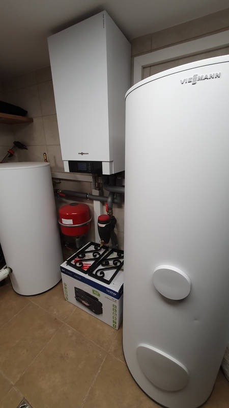 Biały, wiszący piec gazowy Viessmann z podłączonymi rurami, obok pionowy, cylindryczny zasobnik ciepłej wody, na podłodze kartonowe pudełko z kuchenką gazową.